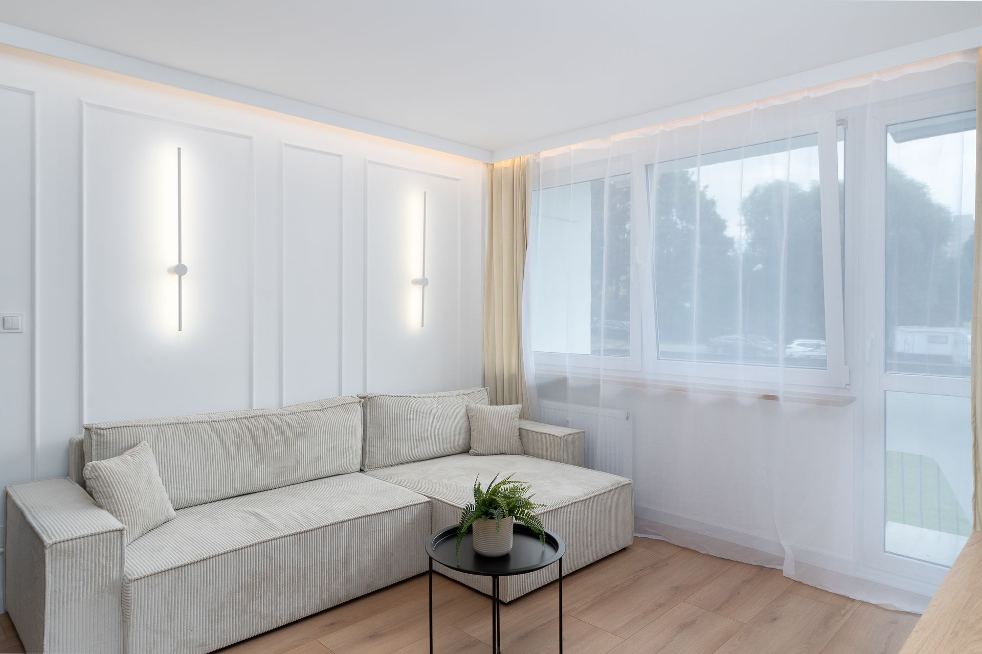 Salon lumineux aux murs blancs, canapé clair et voilages devant une grande fenêtre